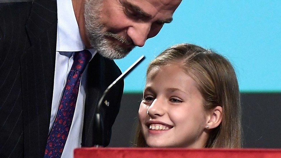 The King of Spain smiles at his daughter, Princess Leonor, as she peeks over the top of a podium almost as tall as she is