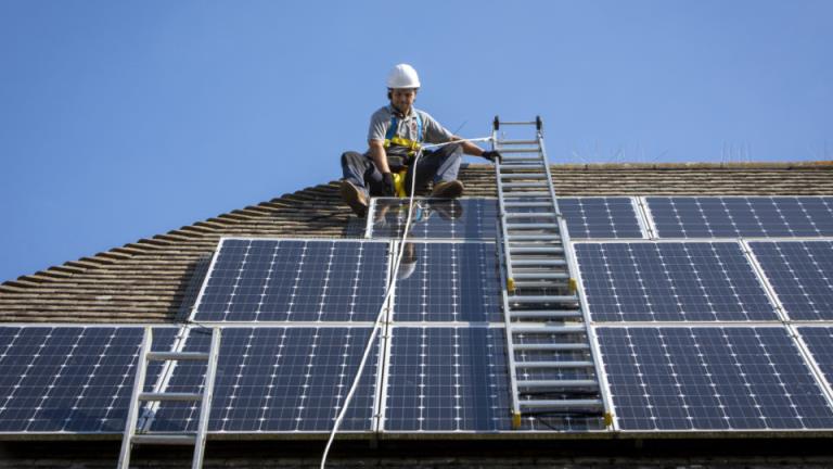 A maintenance person uses a ladder and harnesses to install equipment around a Solar panel array on the roof of a house